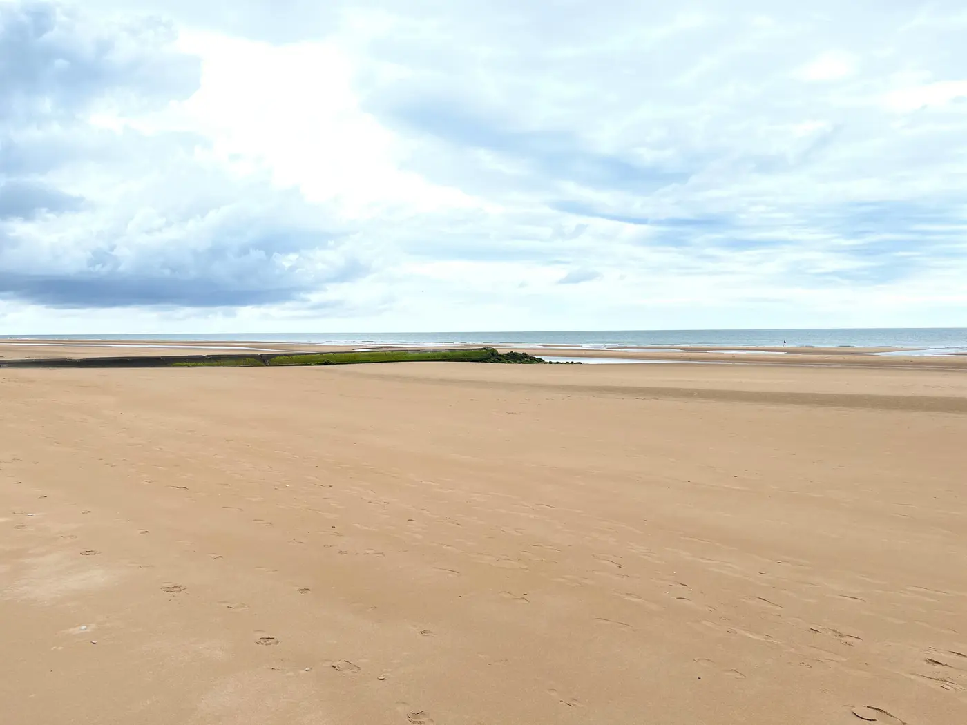 A wide, flat, empty stretch of sand at Omaha Beach with calm water and a pale sky.