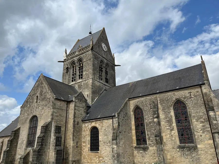 The stone church at Sainte-Mère-Église under a bright sky, with a parachute and effigy hanging from the tower.
